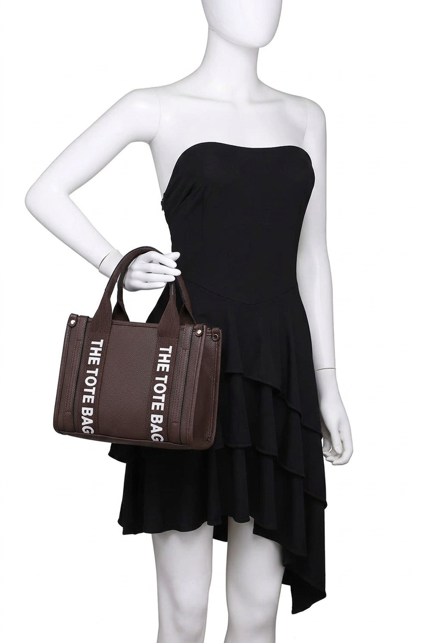 A brown tote bag with white 'THE TOTE BAG' text printed vertically, held by a white mannequin wearing a black strapless ruffled dress against a white background.