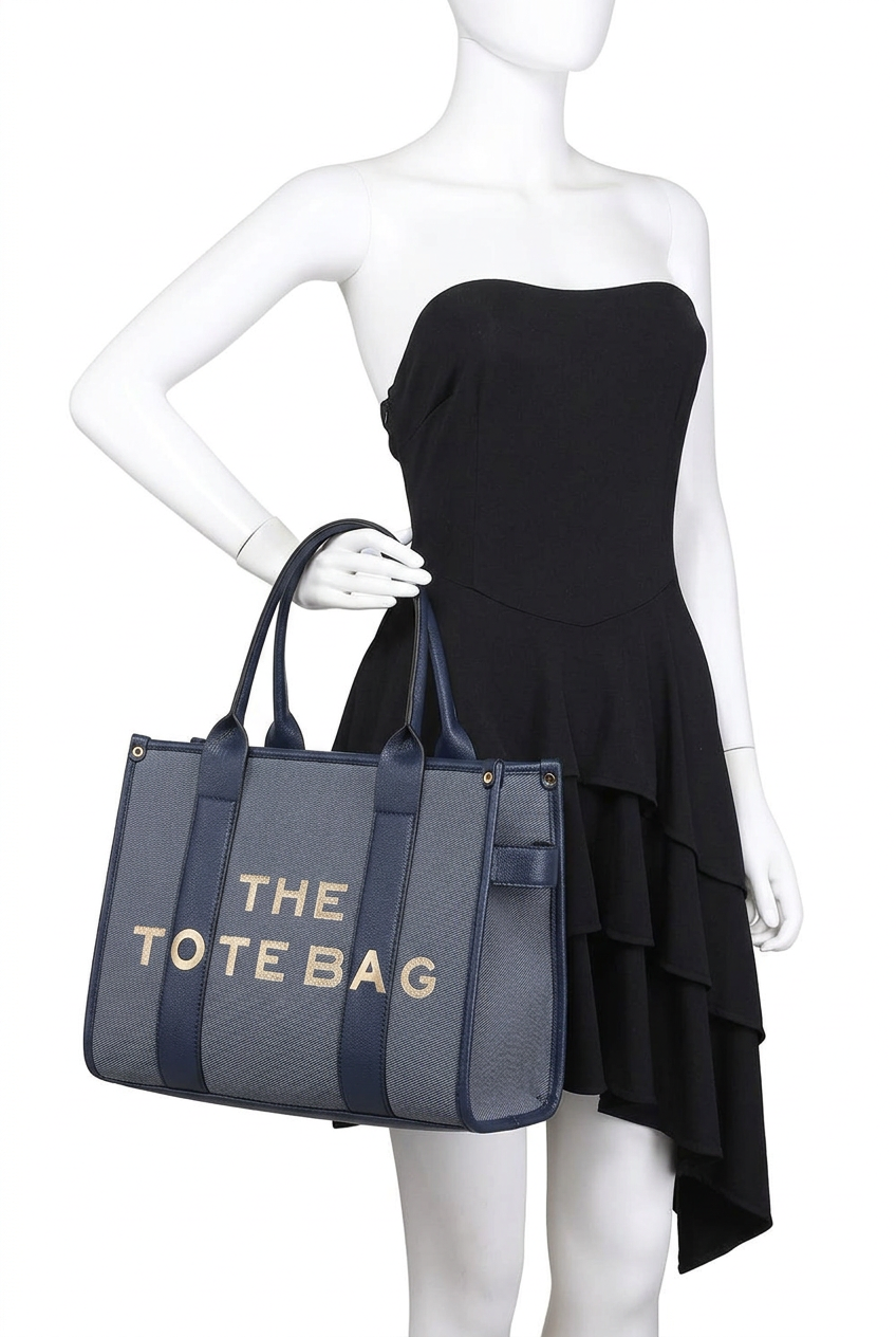 A mannequin wearing a black strapless dress and holding a large tote bag. The tote bag is gray with dark blue handles and side panels, and features the text 'THE TOTE BAG' in gold lettering on the front.