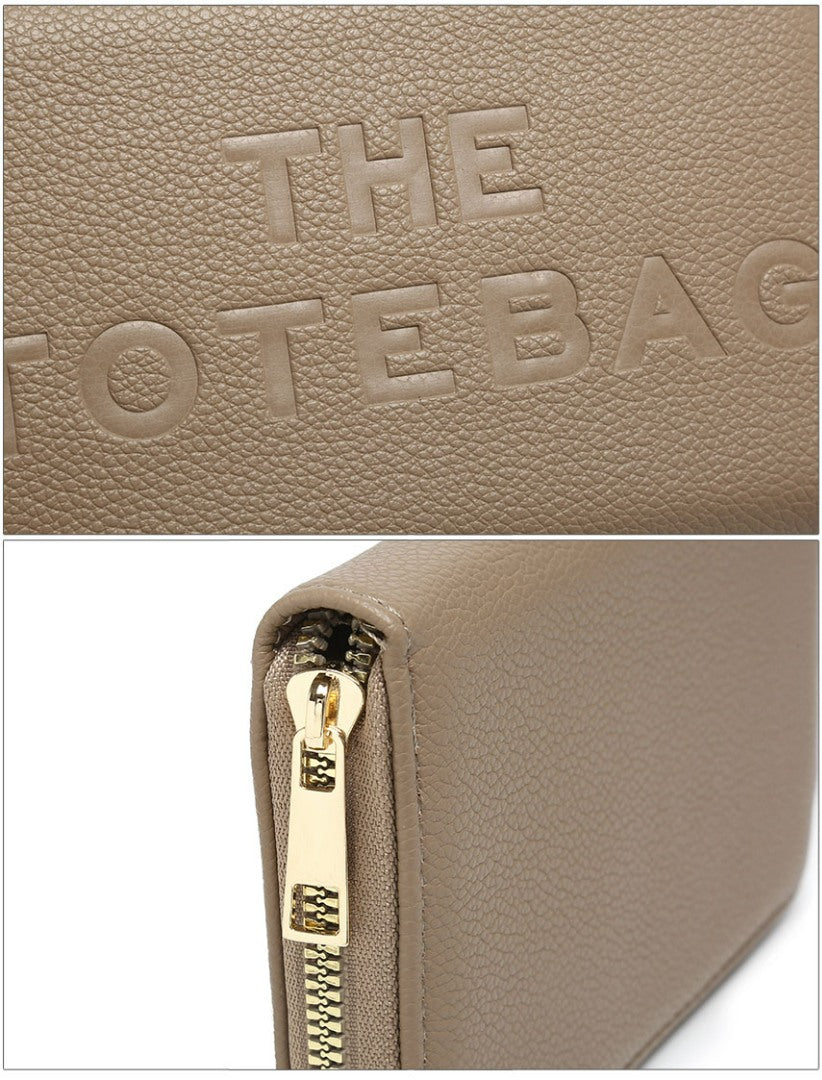 Close-up of a taupe pebbled wallet or clutch with embossed 'THE TOTE BAG' branding on the top panel and a gold-tone zipper pull shown in the lower section.