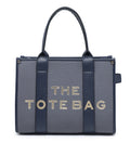 A structured tote bag by The Tote Bag featuring a textured slate blue-gray body with dark navy blue handles and side panels, and 'THE TOTE BAG' printed in metallic gold lettering on the front.