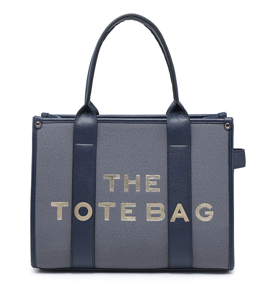 A structured tote bag by The Tote Bag featuring a textured slate blue-gray body with dark navy blue handles and side panels, and 'THE TOTE BAG' printed in metallic gold lettering on the front.