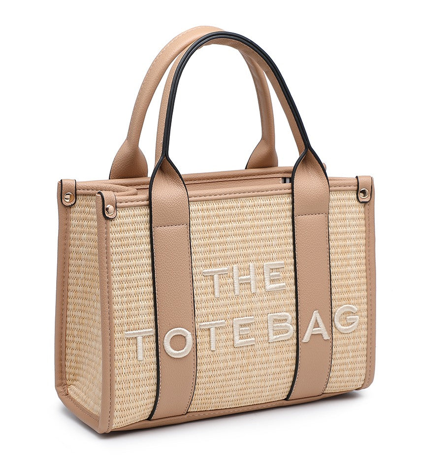 A woven straw tote bag with beige faux-leather trim and handles, featuring the brand name 'THE TOTE BAG' in white lettering on the front, set against a plain white background.
