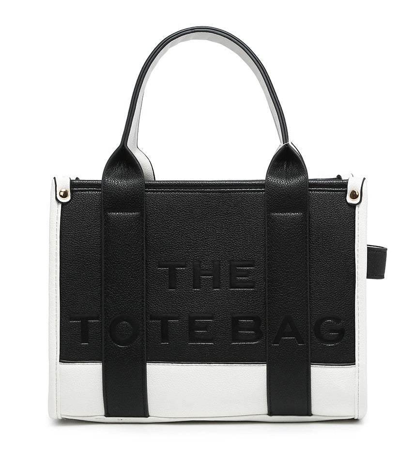 A black and white tote bag with embossed 'THE TOTE BAG' branding on the front, featuring dual top handles and a structured rectangular shape, shot against a plain white background.