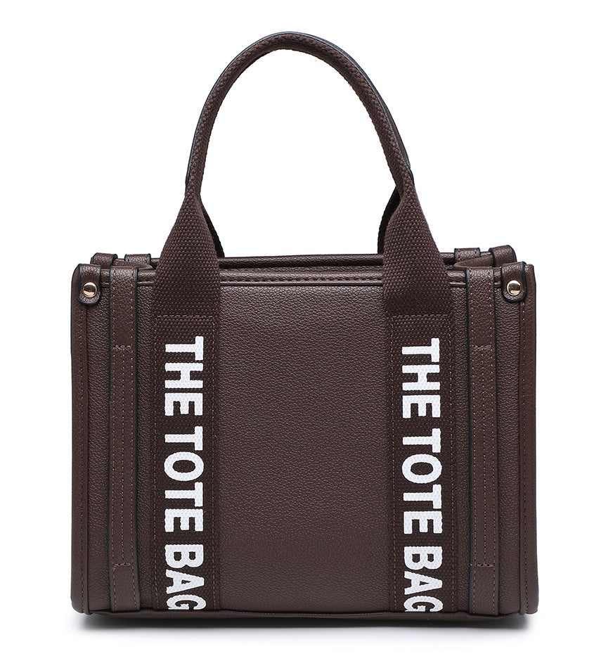 A dark brown pebbled tote bag with dual top handles and bold white vertical text reading 'THE TOTE BAG' on both sides, shown against a plain white background.