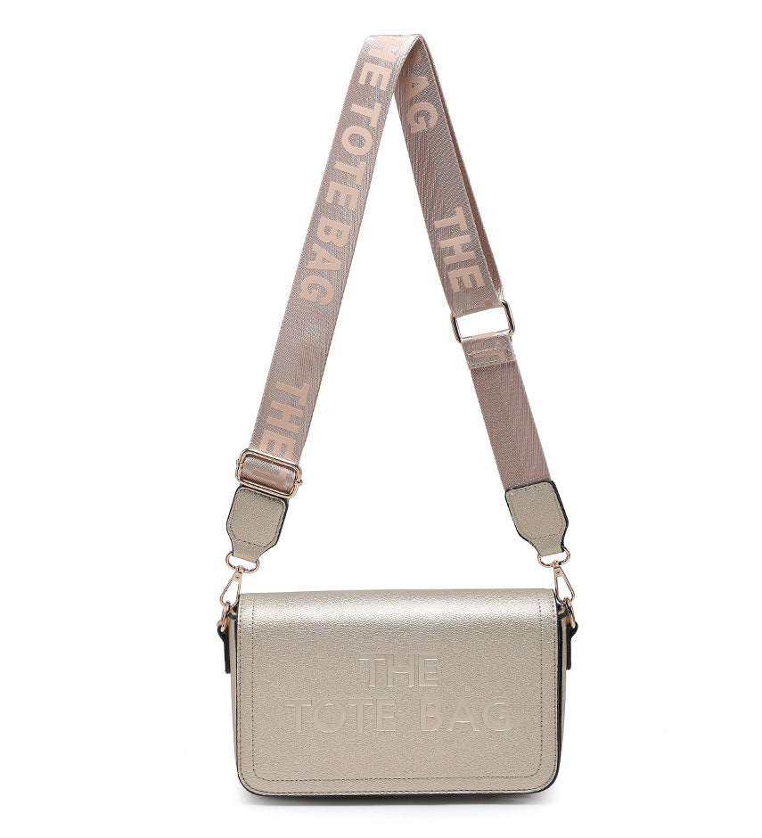 A metallic gold crossbody bag with a wide, adjustable taupe strap featuring the text 'THE TOTE BAG' in beige. The front of the bag has 'THE TOTE BAG' debossed in a matching metallic silver tone.