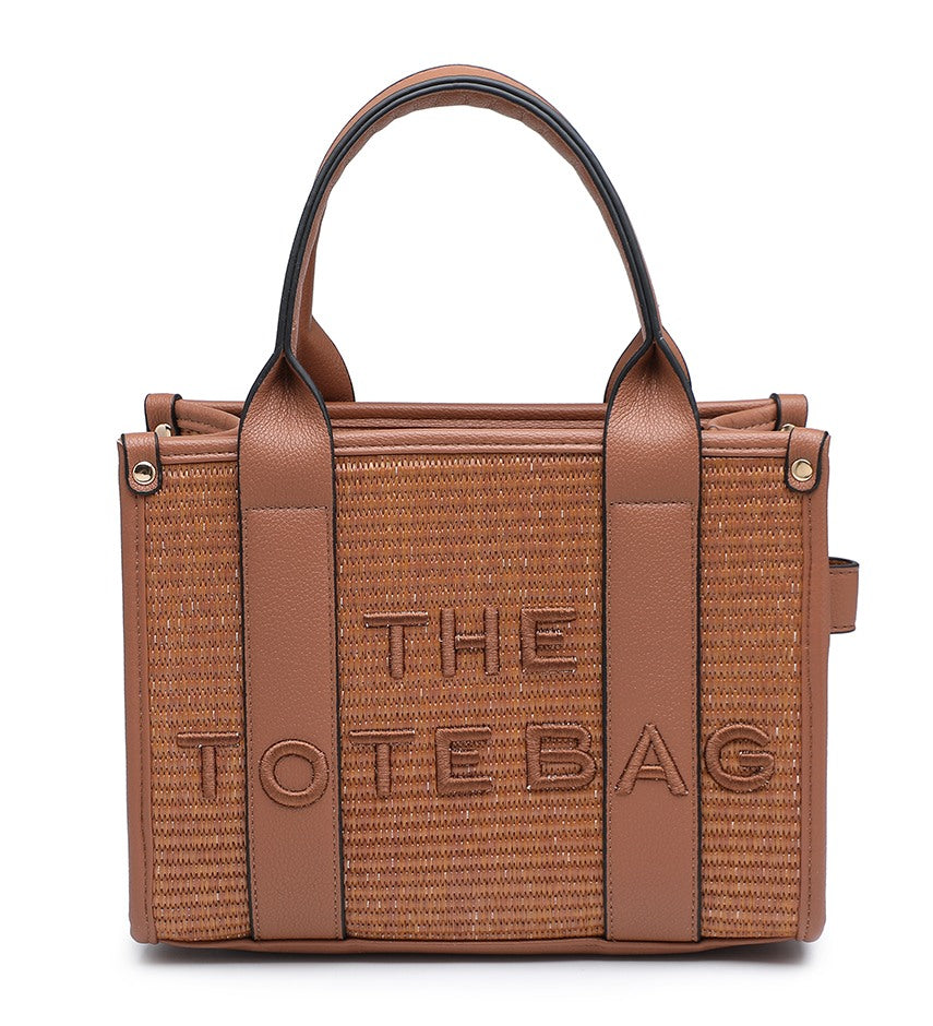 A structured brown tote bag with a woven straw-textured body, smooth brown leather trim and handles, and the words 'THE TOTE BAG' embossed on the front, shown on a plain white background.