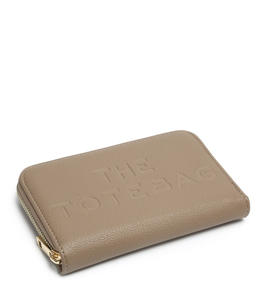 A taupe zip-around tote bag with 'THE TOTE BAG' embossed on the front, featuring a gold-tone zipper pull, shown on a plain white background.