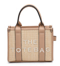A structured woven tote bag in beige and tan with light brown leather handles and trim, featuring the text 'THE TOTE BAG' in white on the front, set against a plain white background.
