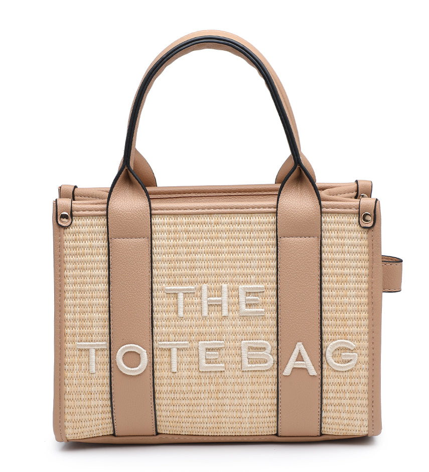 A structured woven tote bag in beige and tan with light brown leather handles and trim, featuring the text 'THE TOTE BAG' in white on the front, set against a plain white background.