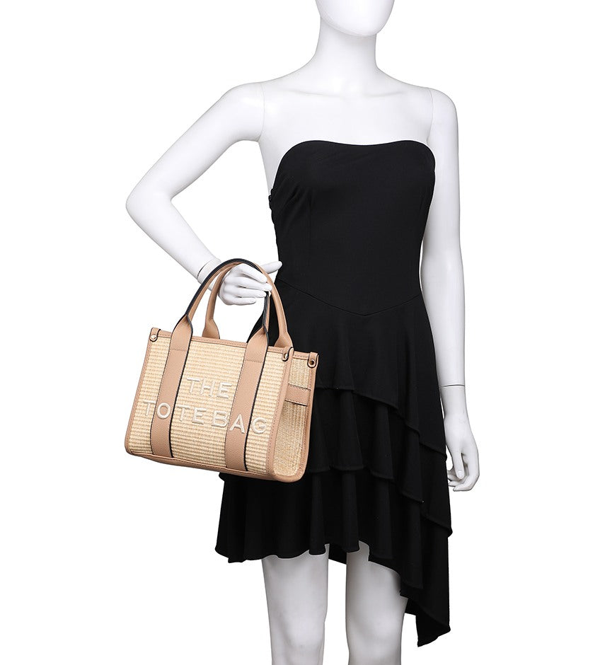 A white mannequin wearing a strapless black dress and holding a beige woven tote bag with tan handles and trim. The bag has large metallic lettering that reads 'THE TOTE BAG'.