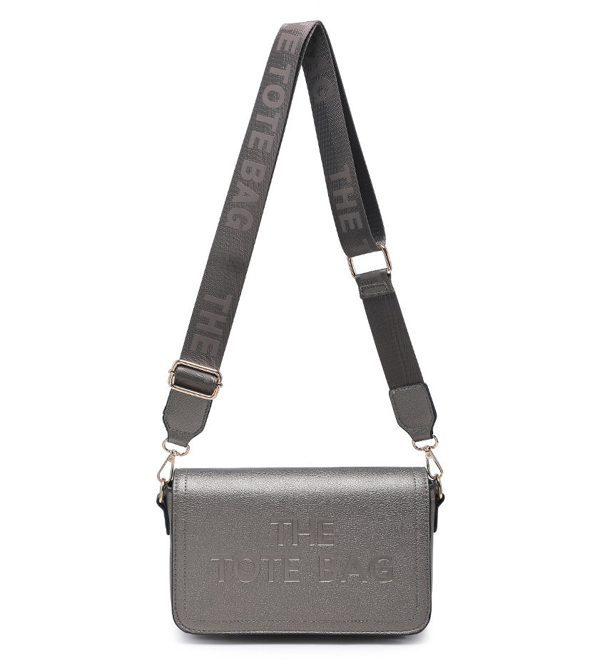 A metallic gray crossbody bag with a matching gray adjustable shoulder strap featuring the text 'THE TOTE BAG' in white. The bag has a textured metallic finish and gold-tone hardware, shown against a plain white background.