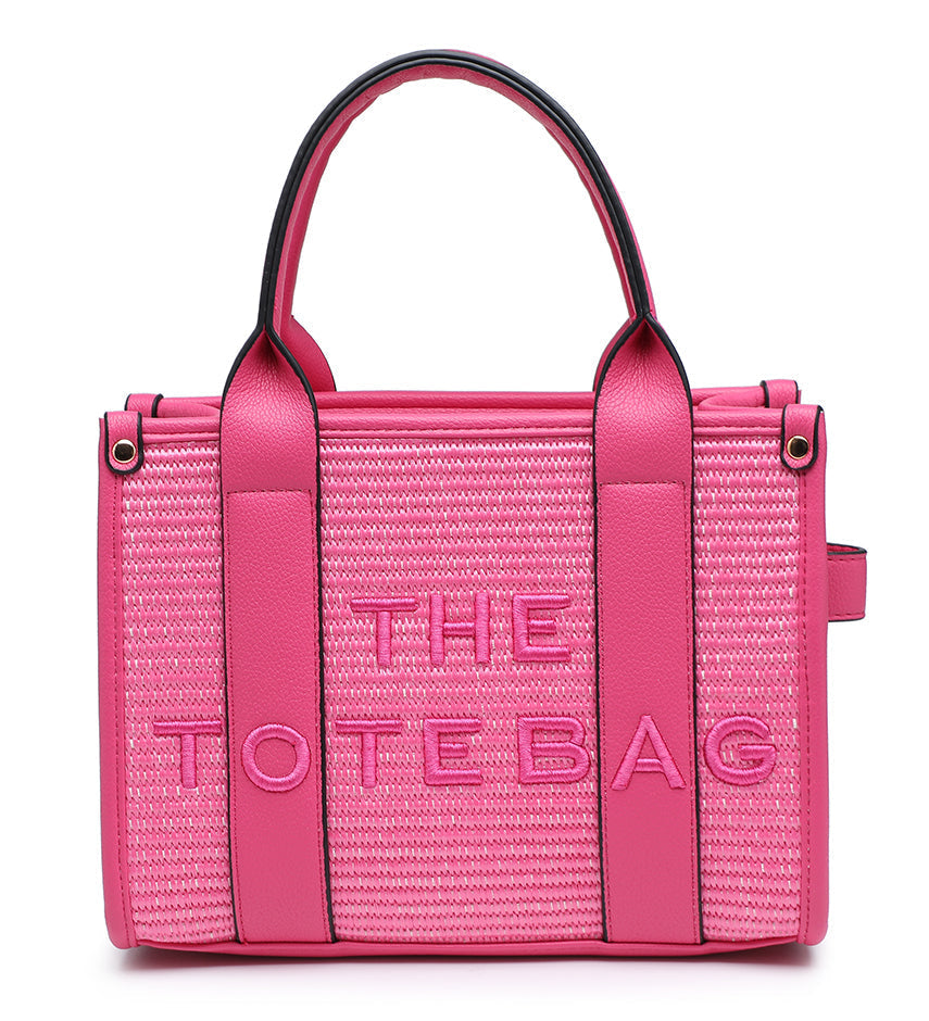 A bright pink woven tote bag with smooth pink leather handles and trim, featuring the text 'THE TOTE BAG' embossed on the front, set against a plain white background.