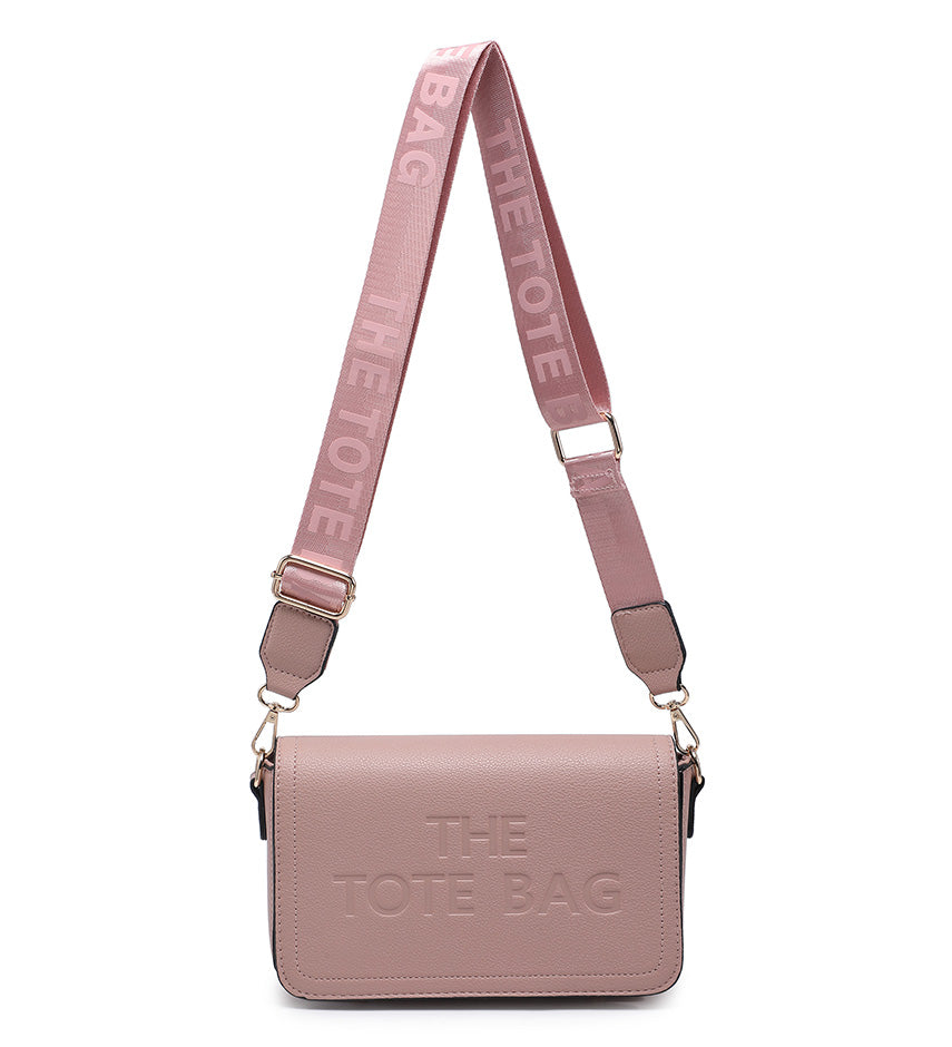 A dusty pink leather shoulder bag with a matching adjustable webbed strap. The bag features the text 'THE TOTE BAG' embossed on the front and printed on the strap, with gold-tone hardware.